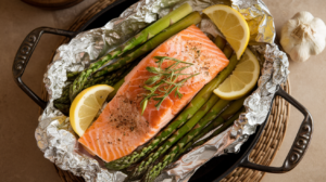 garlic butter baked salmon with lemon & asparagus , featuring tender salmon fillets nestled alongside vibrant asparagus spears, all drizzled with a golden garlic butter sauce and garnished with fresh lemon wedges and herbs. The dish is baked in aluminum foil , creating a moist and flavorful cooking environment, and served hot for an inviting and appetizing presentation.