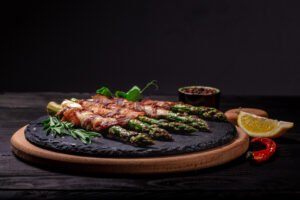 A beautifully arranged plate of prosciutto-wrapped asparagus , featuring tender green spears tightly wrapped in golden-brown prosciutto slices. The dish is garnished with fresh herbs and served on a rustic wooden board, making it an inviting and festive addition to any meal.